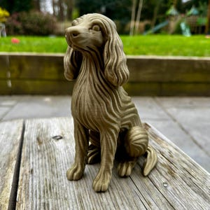 May include: A detailed, seated dog sculpture in a light brown color. The dog has long, wavy fur and is positioned on a wooden surface. The sculpture is a decorative item, likely for home decor.