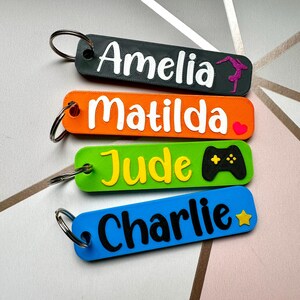 Puede incluir: Cuatro llaveros coloridos con nombres: Amelia, Matilda, Jude y Charlie. Cada llavero tiene un color de fondo diferente: negro, naranja, verde y azul. Los nombres son en blanco, con un peque&ntilde;o gr&aacute;fico: una gimnasta, un coraz&oacute;n, un mando de videojuegos y una estrella.
