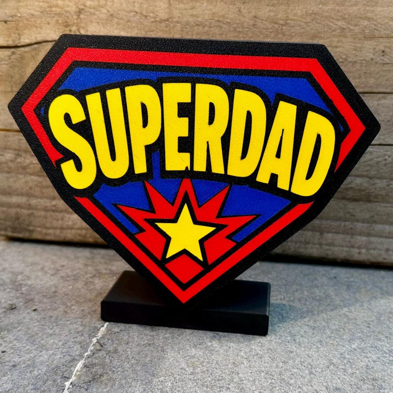 May include: A superhero-themed sign with the word "SUPERDAD" in yellow, set against a blue background within a red-bordered shield shape. A yellow star is at the bottom. The sign is mounted on a black base.
