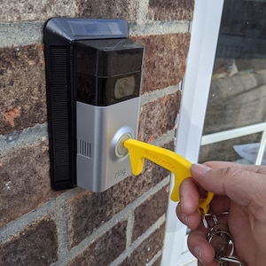 May include: A yellow door opener tool with the text "Wow London" printed on it. The tool is being used to open a Ring doorbell.