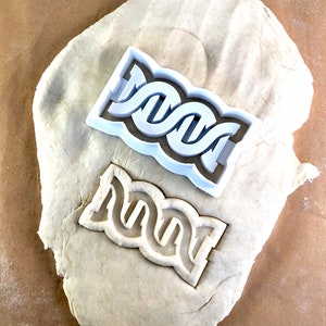 Könnte beinhalten: Ein weißer Plastik-Ausstecher in Form einer Doppelhelix-DNA-Strang. Der Ausstecher liegt auf einer weißen Oberfläche, mit einem zweiten Abdruck des Ausstechers in der Oberfläche.