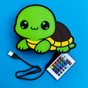 K&ouml;nnte beinhalten: Gr&uuml;ne, schildkr&ouml;tenf&ouml;rmige LED-Lampe mit braunem Panzer und gelben Akzenten. Die Lampe enth&auml;lt ein USB-Kabel und eine Fernbedienung mit Farboptionen und einem Ein/Aus-Schalter. Der Hintergrund ist einfarbig blau.