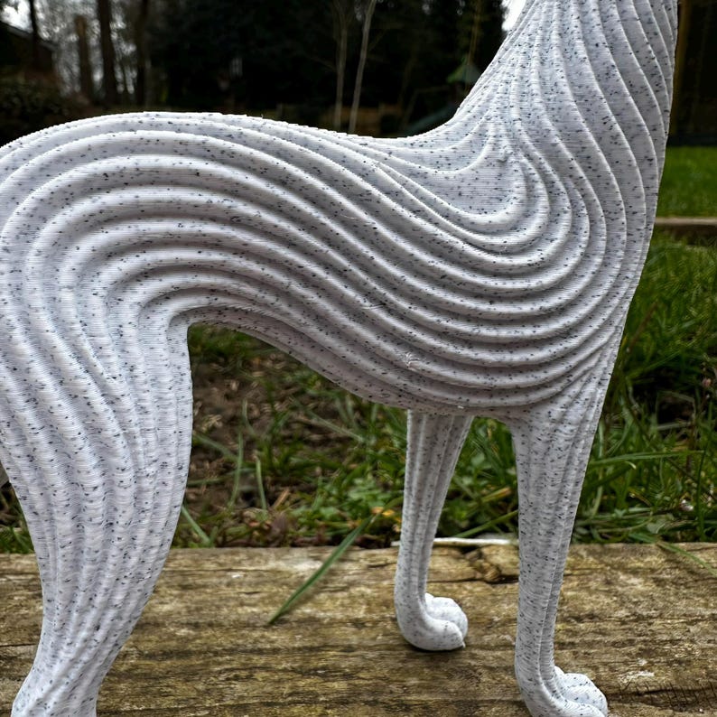 May include: A white and gray speckled sculpture of a dog, with a streamlined, modern design. The dog's body features a series of curved, wave-like ridges. The sculpture is positioned on a wooden surface, with a blurred green background.