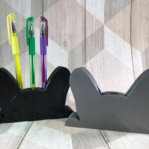 May include: Two 3D printed pen holders shaped like a French bulldog. The black holder has three pens in it: a yellow pen, a green pen, and a purple pen. The gray holder is empty.