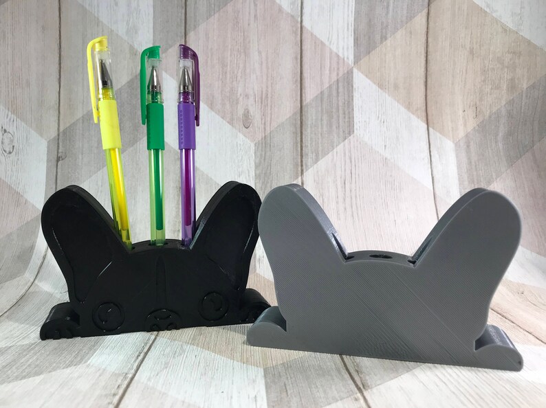 May include: Two 3D printed pen holders shaped like a French bulldog. The black holder has three pens in it: a yellow pen, a green pen, and a purple pen. The gray holder is empty.