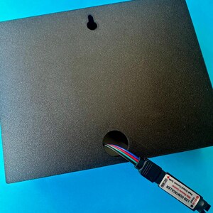 May include: A black rectangular LED controller box with a textured surface. A hole is cut out of the box to allow a multi-coloured wire to pass through. The box has a small hole for hanging.