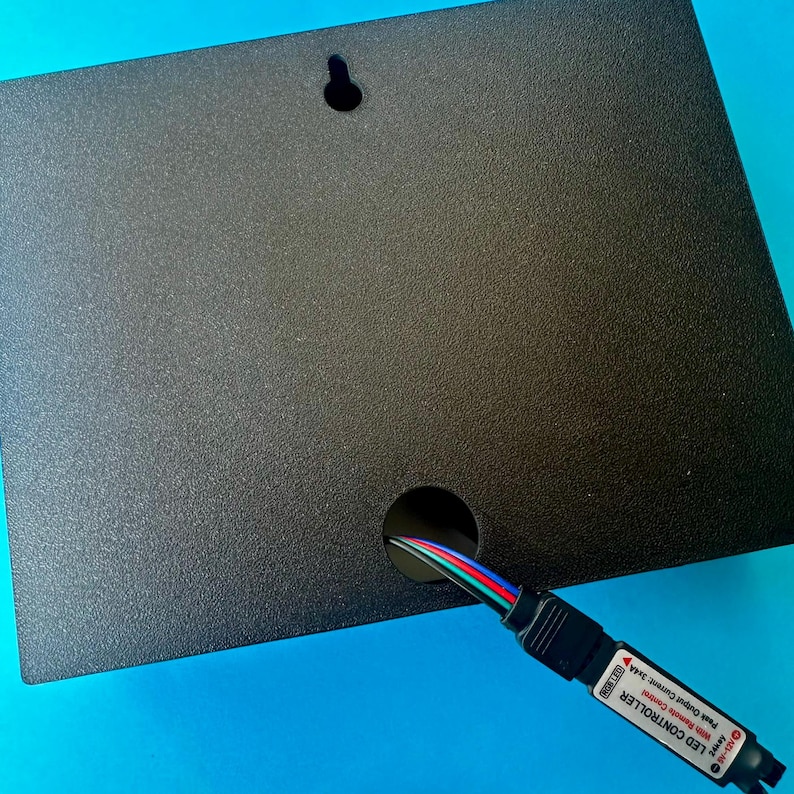 May include: A black rectangular LED controller box with a textured surface. A hole is cut out of the box to allow a multi-coloured wire to pass through. The box has a small hole for hanging.