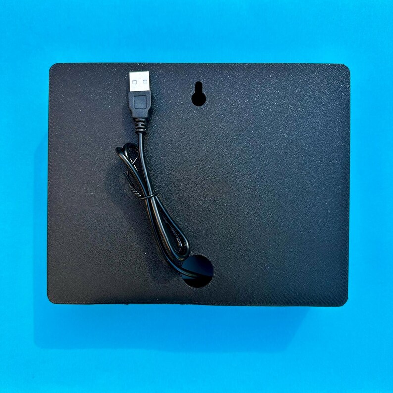 May include: A black rectangular panel with a textured surface, a USB cable, and a small hole. The USB cable is connected to the panel and has a USB-A connector. A small hole and a keyhole-shaped hanger are visible.