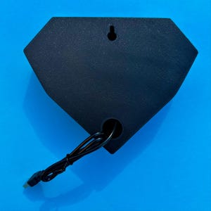 May include: A black, diamond-shaped object with a textured surface. It has a hole at the bottom and a small hole at the top for hanging. A black wire with a plug extends from the bottom. The object is set against a bright blue background.