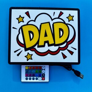 May include: A rectangular sign with the word "DAD" in yellow, outlined in black and red, in a comic book style. The sign has a black border, star accents, and a remote control with color options. A USB cable is attached.