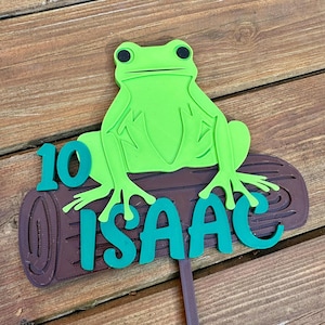 May include: A green frog sits atop a brown log, with the name "ISAAC" and the number "10" in teal. The frog has black eyes and is a vibrant green color. The log has a textured appearance. The item is likely a cake topper.