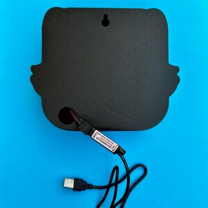 May include: Black, wall-mounted LED light with a unique shape. The back features a hole for the power cord and a small hole for hanging. A USB cable and LED controller are attached. The background is a solid blue.