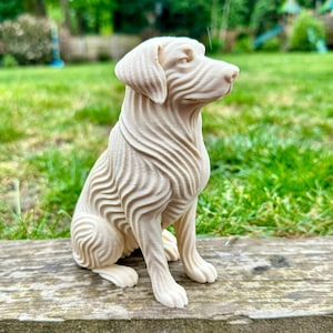 May include: A cream-colored, seated dog sculpture with a unique, textured design. The dog's fur is depicted with flowing, wave-like lines. The sculpture is placed on a wooden surface against a backdrop of green grass and foliage.
