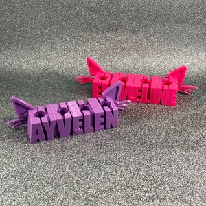 May include: Two purple and pink cat-shaped pencil holders with the names "Ayvellen" and "Emaline" spelled out in raised letters.