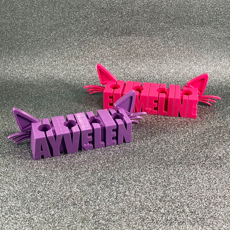 May include: Two purple and pink cat-shaped pencil holders with the names "Ayvellen" and "Emaline" spelled out in raised letters.