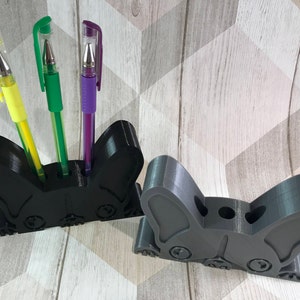 May include: Two black and gray 3D printed pen holders shaped like a French bulldog. The holder on the left has three pens in it: a yellow pen, a green pen, and a purple pen.
