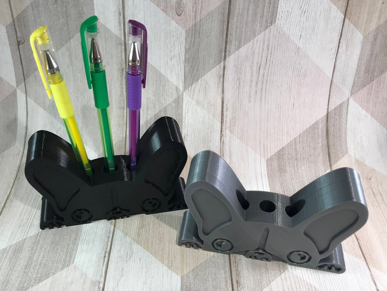 May include: Two black and gray 3D printed pen holders shaped like a French bulldog. The holder on the left has three pens in it: a yellow pen, a green pen, and a purple pen.