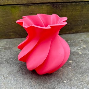 May include: A vibrant, hot pink vase with a unique, swirling design. The vase has a rounded base that tapers upwards, with petal-like curves creating its form. The top edge has a ruffled appearance. The vase is made of a smooth material.