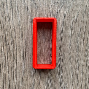 May include: A bright red plastic frame with a rectangular opening. The frame is on a woodgrain surface.