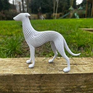 May include: A gray, stylized sculpture of a greyhound dog. The dog is standing on a wooden surface, with a green grassy background. The sculpture has a unique design with horizontal wave-like lines across its body.