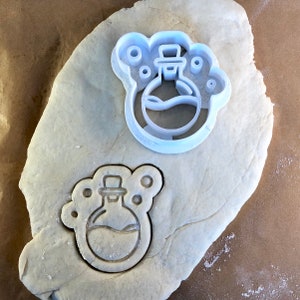 Puede incluir: Un cortador de galletas de plástico blanco con forma de botella de poción con burbujas alrededor. El cortador de galletas está sobre una superficie blanca.