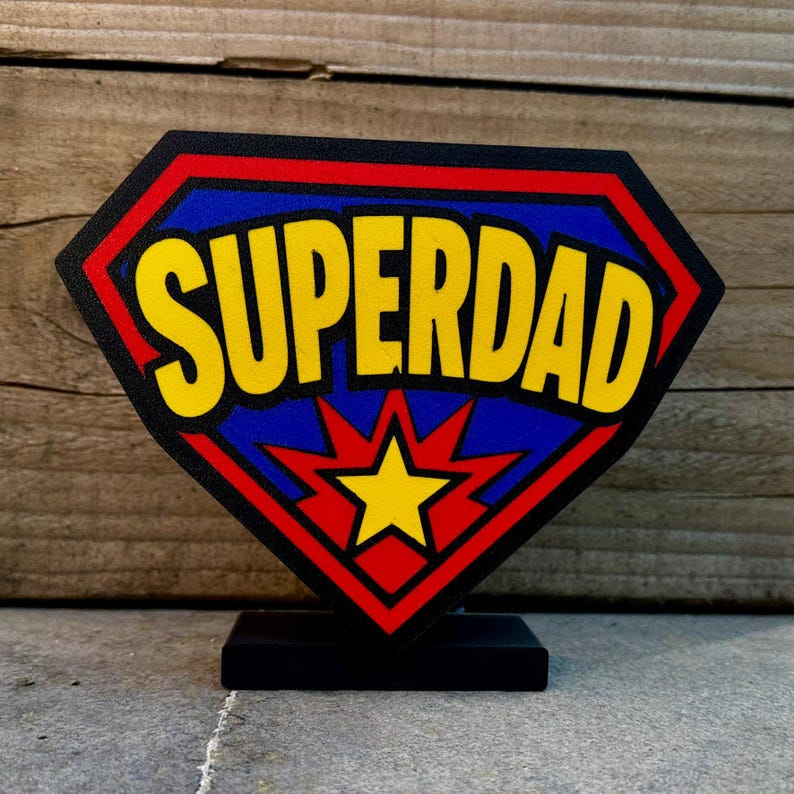 May include: A black diamond-shaped sign with the word "SUPERDAD" in yellow, outlined in black, set against a blue background with a red border. A yellow star is at the bottom. The sign is mounted on a black base.