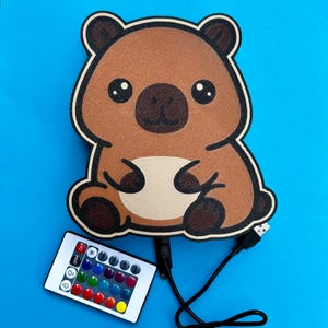 May include: A brown bear-shaped LED night light with a black outline, sitting pose, and a remote control. The remote has multiple color options. The night light has a USB cable for power.