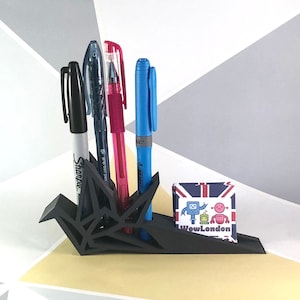 May include: Black origami paper crane desk organiser with four pens, including a blue pen, a pink pen, a black pen, and a white pen with the word "Sharpie" on it. A small card with the text "WowLondon" and a cartoon robot design is in front of the organiser.