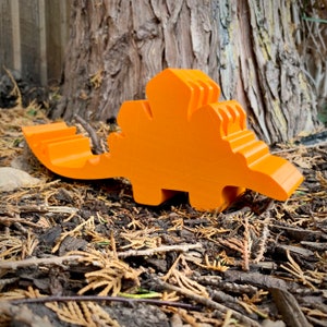 May include: Orange 3D printed dinosaur toy. The dinosaur is standing on a bed of brown pine needles.