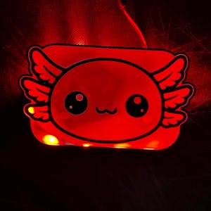 K&ouml;nnte beinhalten: Ein rotes, leuchtendes Nachtlicht in Axolotl-Form. Das Licht hat ein cartoonartiges Design mit gro&szlig;en Augen und fl&uuml;gelartigen Merkmalen. Das Licht ist eingesteckt und strahlt ein weiches, rotes Leuchten aus.
