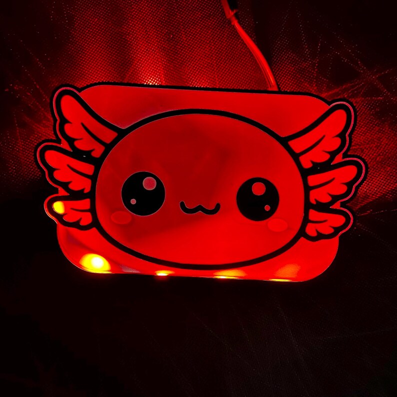 K&ouml;nnte beinhalten: Ein rotes, leuchtendes Nachtlicht in Axolotl-Form. Das Licht hat ein cartoonartiges Design mit gro&szlig;en Augen und fl&uuml;gelartigen Merkmalen. Das Licht ist eingesteckt und strahlt ein weiches, rotes Leuchten aus.