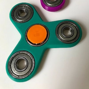May include: A teal green fidget spinner with three metal bearings and an orange centre cap.