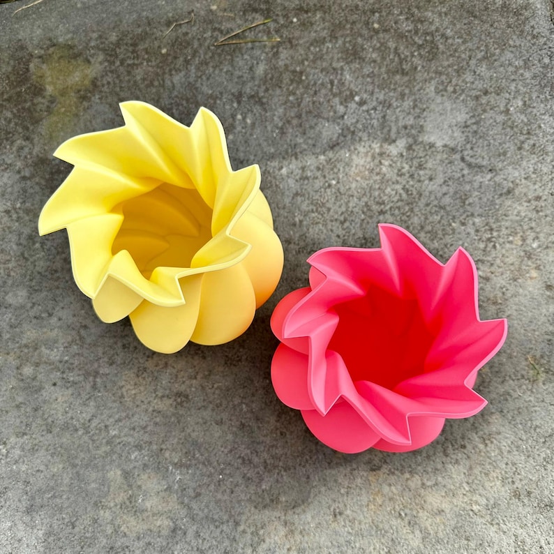 May include: Two flower-shaped containers, one yellow and one pink, sit on a gray surface. The containers have a layered petal design, with the yellow one on the left and the pink one on the right. The containers are empty.