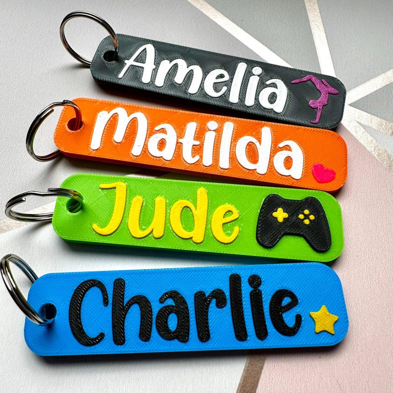 Puede incluir: Cuatro llaveros personalizados en varios colores: gris oscuro, naranja, verde y azul. Cada llavero presenta un nombre en un color contrastante, con peque&ntilde;os iconos como una gimnasta, un coraz&oacute;n, un mando de juegos y una estrella.
