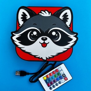 May include: A raccoon-shaped LED light with a red background and a blue background. The raccoon has black and white markings and a smiling expression. A remote control and USB cable are included.