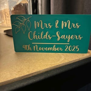 Op de afbeelding: Teal trouwteken met gouden letters. Het bord leest "Mrs & Mrs Childs-Sayers 4th November 2025" met een bladerontwerp. Het bord is gemaakt van een stevig materiaal en staat op een lichtbruin oppervlak.