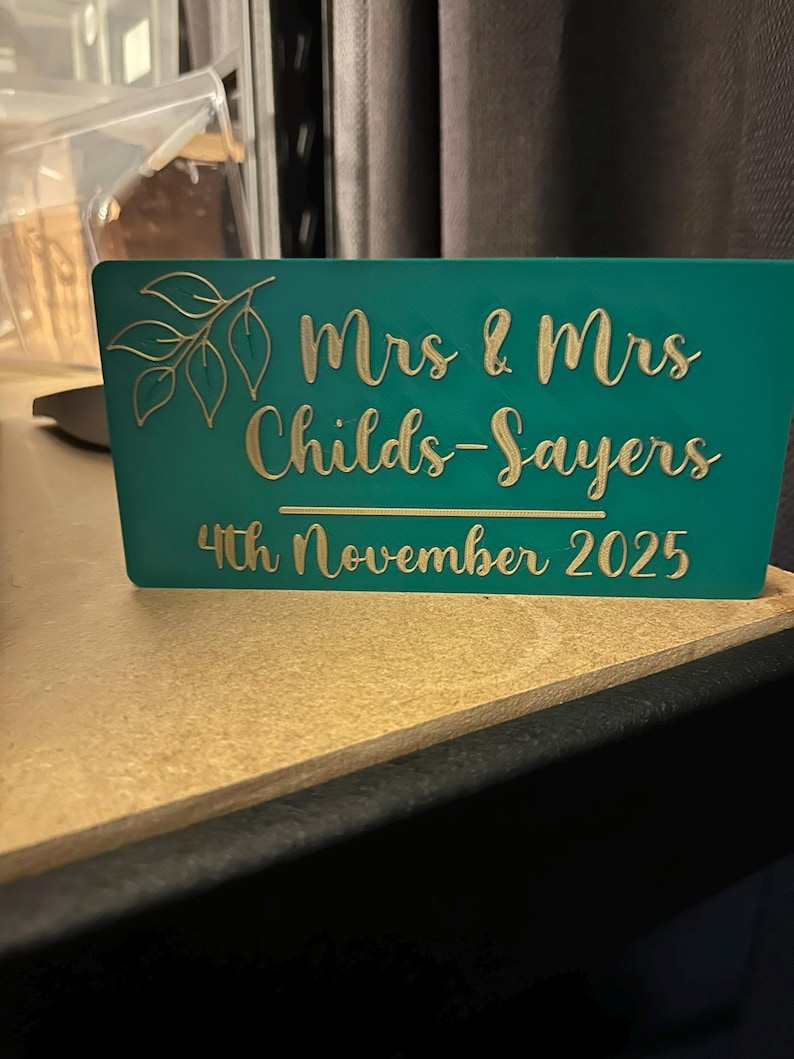 Op de afbeelding: Teal trouwteken met gouden letters. Het bord leest "Mrs & Mrs Childs-Sayers 4th November 2025" met een bladerontwerp. Het bord is gemaakt van een stevig materiaal en staat op een lichtbruin oppervlak.