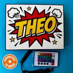 May include: A personalised light-up sign featuring the name "THEO" in yellow, outlined in black, set against a red burst design. Includes a remote control with colour options and a USB cable. The sign is white with black trim and has a comic book style.