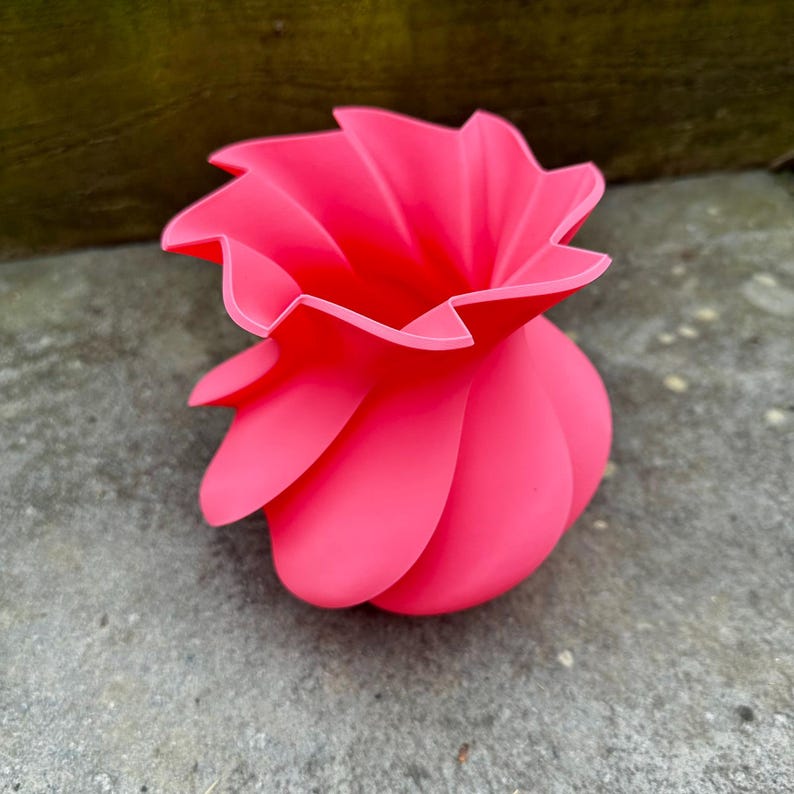 May include: A vibrant pink, abstract flower-shaped vase. The vase has a unique design with petal-like curves that spiral upwards. The bright color and unusual shape make it a striking decorative piece.