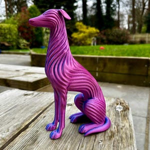 May include: A magenta and blue decorative dog sculpture. The dog is sitting with its legs tucked beneath it. The sculpture has a striped pattern and is made of a solid material. The dog has a long neck and pointed ears. The sculpture is sitting on a wooden surface.
