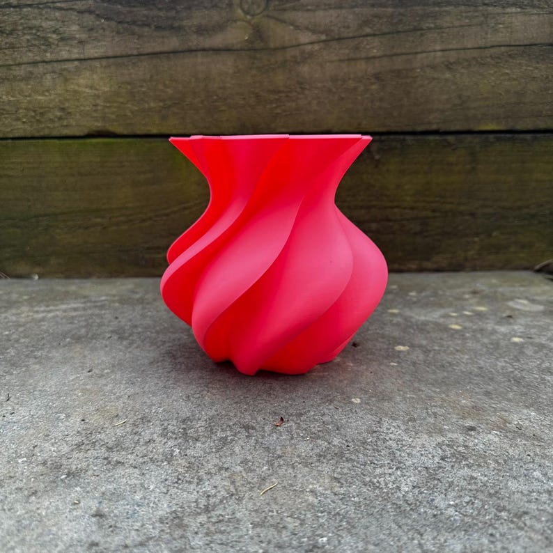 May include: A vibrant red vase with a unique, swirling design. The vase has a bulbous base and a flared top, with the body of the vase featuring a series of curved, petal-like shapes. The vase is made of a smooth material and sits on a concrete surface.