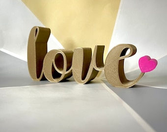 May include: A gold-colored, 3D-printed "love" sign with a pink heart attached to the letter "e". The sign is placed on a surface with geometric shapes in gold, white, and gray.