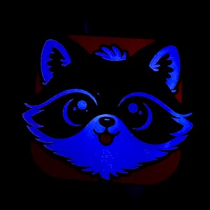 May include: A raccoon face design with blue neon outlines and accents against a black background. The raccoon's eyes, nose, and mouth are detailed with blue light. The overall design is a cartoon style.