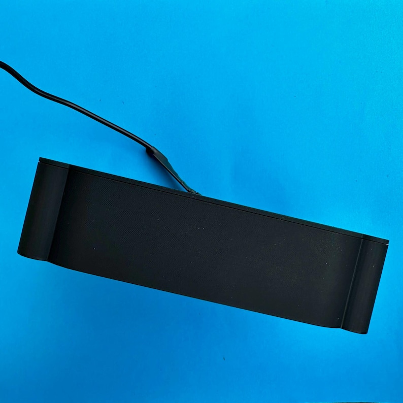 K&ouml;nnte beinhalten: Ein schwarzes rechteckiges elektronisches Ger&auml;t mit abgerundeter Kante und einem schwarzen Kabel, das sich von oben erstreckt. Das Ger&auml;t befindet sich vor einem leuchtend blauen Hintergrund.