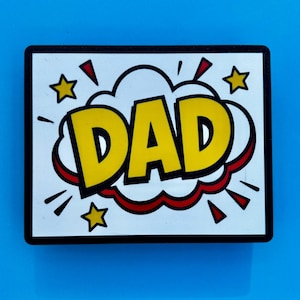 May include: A rectangular sticker with a white background and a black border. The word "DAD" is in yellow, inside a white speech bubble with red and black accents. Yellow stars and black lines complete the design.