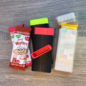May include: Three pouches of baby food, two are black with red and yellow accents and one is clear with a white label that says "Kiddylicious". The pouches are labeled "Carrot Wafers" and are gluten and lactose free.