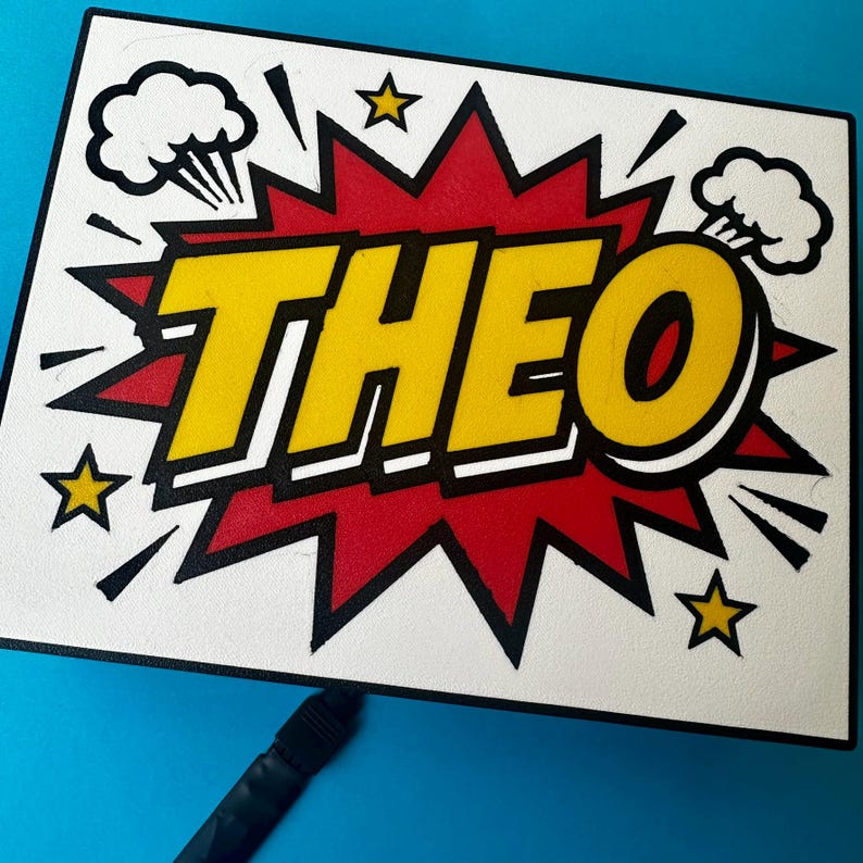 May include: A rectangular sign featuring the name "THEO" in bold yellow lettering with a black outline, set against a red and white comic book-style backdrop. The design incorporates star and explosion graphics.