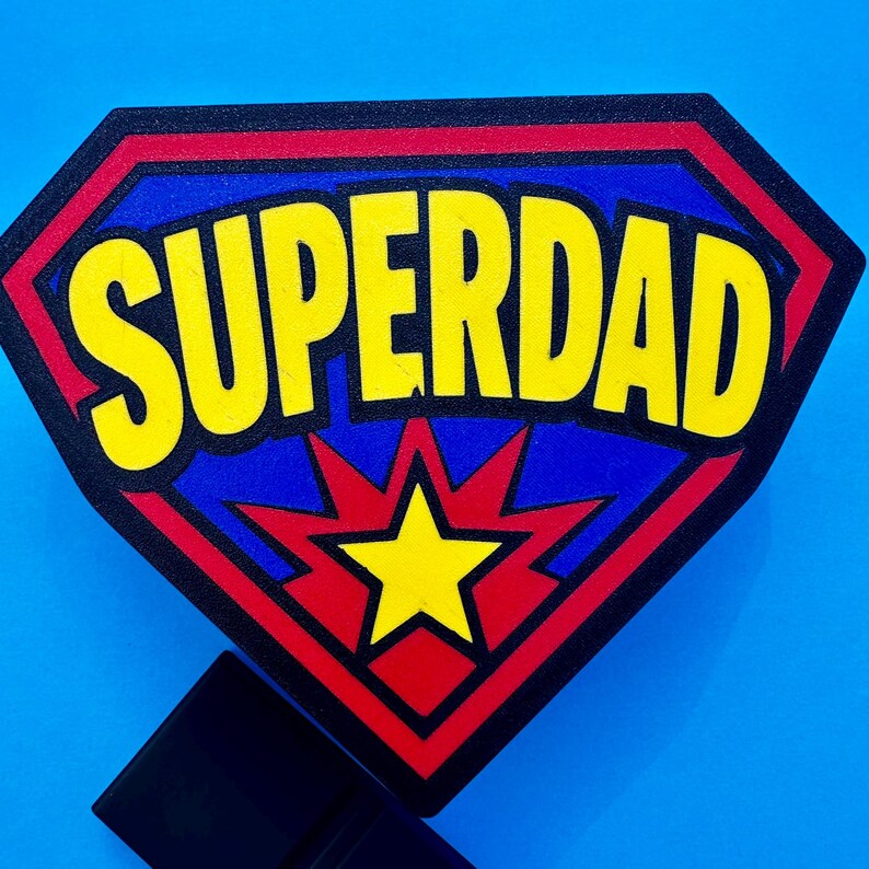 May include: A shield-shaped emblem with the word "SUPERDAD" in yellow, outlined in black, set against a blue background. The emblem has a red border and a yellow starburst design at the bottom. A black stand is attached.