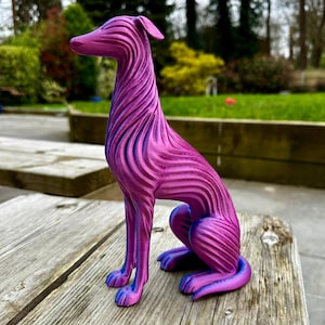 May include: A decorative sculpture of a dog in a seated position. The dog is primarily magenta with blue accents, featuring a striped pattern across its body. The sculpture is placed on a weathered wooden surface, with a blurred green background.