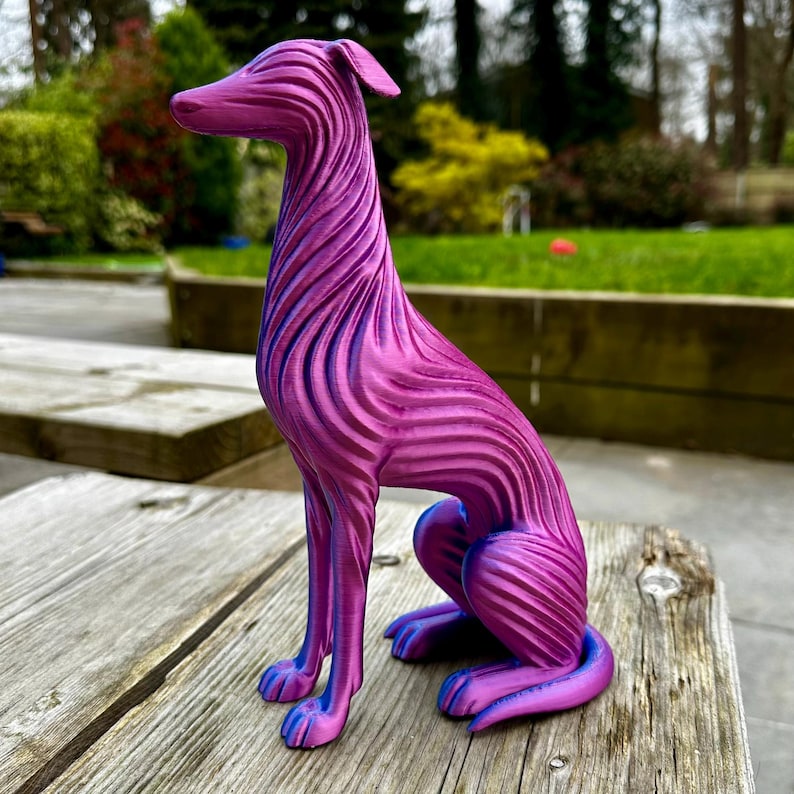 May include: A decorative sculpture of a dog in a seated position. The dog is primarily magenta with blue accents, featuring a striped pattern across its body. The sculpture is placed on a weathered wooden surface, with a blurred green background.